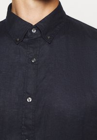 Gros plan sur une chemise bleu marine foncé avec un col boutonné, comportant plusieurs boutons le long de la patte de boutonnage, mettant en valeur son tissu texturé.