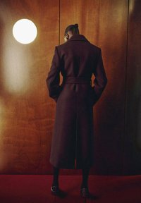 Cappotto lungo di un marrone scuro con colletto alto, vita cintrata e texture liscia, abbinato a scarpe nere, su uno sfondo di legno.