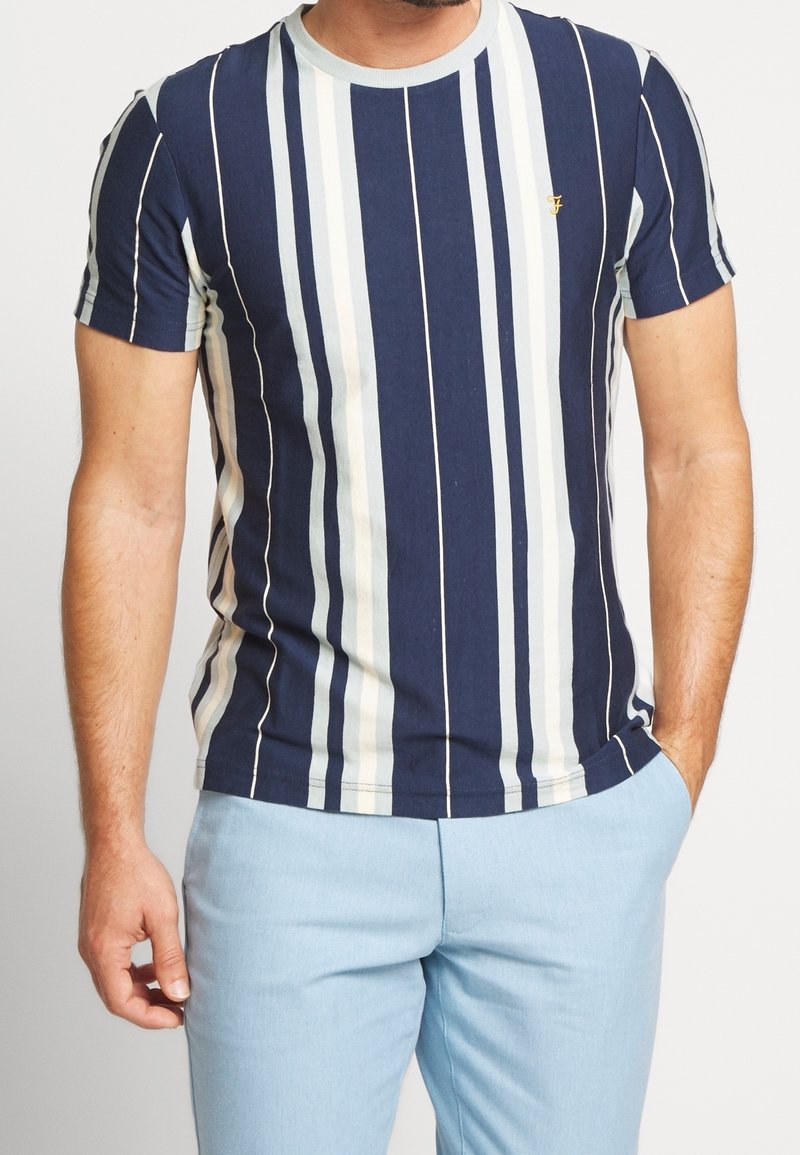 Muž nosící námořnické, bílé a světle modré pruhované tričko s krátkým rukávem a světle modré kalhoty, s jednou rukou v kapse.