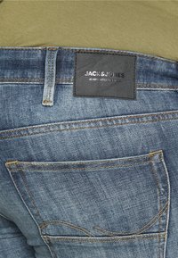 Jeans de mezclilla azul con un acabado texturizado, que presentan costuras naranjas y una etiqueta de cuero negra con "JACK & JONES" en el bolsillo trasero.