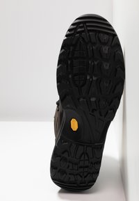 Musta kumipohja, jossa on karhea kuvio, jossa on keltainen Vibram-logo. Kengän sivu on ruskea ja teksturoitu kestävyuden vuoksi.