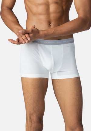 Männlicher Oberkörper trägt weiße Boxershorts mit grauem Bund; die Hände sind vor dem unteren Bauch verschränkt.