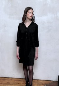 Schwarzes Hemdkleid aus glattem Stoff mit Kragen, halblangen Ärmeln und lockerem Schnitt, das bis zur Mitte des Oberschenkels reicht.