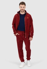 Roter Samt Trainingsanzug mit Reißverschlussjacke und Hose mit elastischem Bund. Enthält ein goldenes Logo auf der Jacke und weiße Sneakers.