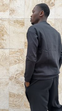 Conjunto de sudadera y pantalones de chándal negros, con un diseño sencillo, puños acanalados y bolsillos laterales, frente a una pared beige texturizada.