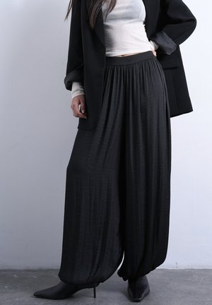 Nő fekete bőszárú nadrágot, fekete blézert, fehér hosszú ujjú felsőt és fekete, hegyes orrú, magassarkú cipőt visel, és egy sima falnak támaszkodik.