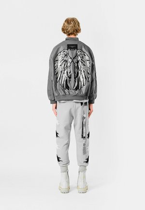 Chaqueta bomber gris con bordado de alas negras y plateadas en la parte posterior, combinada con pantalones de chándal grises con acentos negros desgastados.