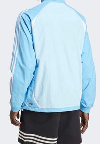 Ljusskär windbreaker-jacka med vita detaljer och tre ränder på ärmarna. Har elastiska manschetter och en avslappnad passform. Tyget är slätt.