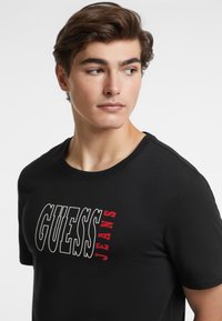 Juoda medvilninė marškinė su „GUESS JEANS“ tekstu ryškiai raudonai ir baltai atspausdintu. Klasikinis apvalus kaklas ir trumpomis rankovėmis.