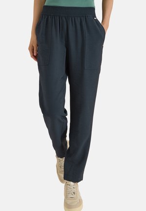 Personne portant un pantalon décontracté bleu foncé avec une taille élastique et des poches latérales, associé à des baskets beige clair.
