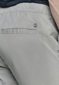 Grijze, lichte shorts met een gladde textuur, voorzien van een achterzak en een hexagonaal logo detail boven de zak.