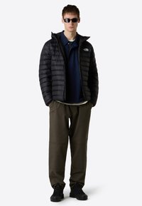Black puffer jacket with horizontal stitching, navy polo shirt, and olive green pants. Black hiking boots complete the outfit. Sunglasses worn.