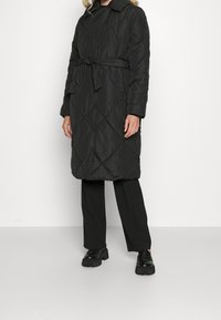 Zwarte gewatteerde lange jas met een ceintuur in de taille, voorzien van een kraag, diagonale patronen en lange mouwen. Gecombineerd met zwarte broek en schoenen.