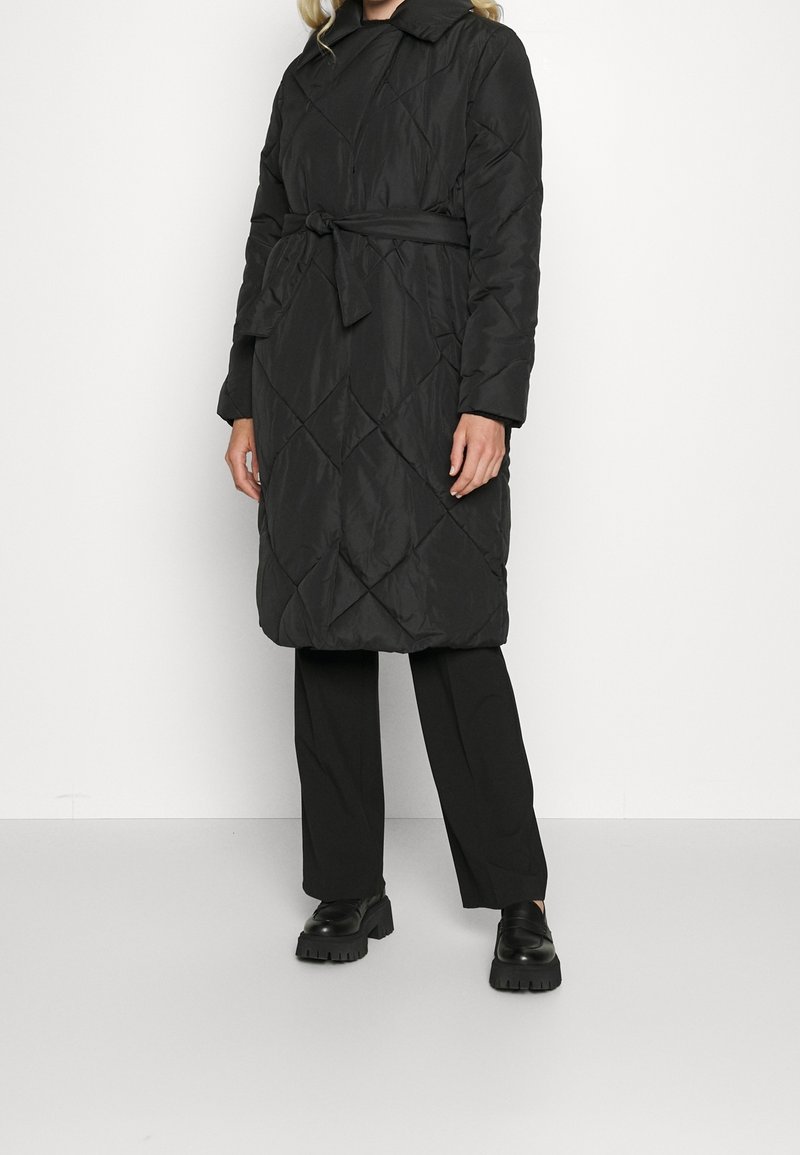 Zwarte gewatteerde lange jas met een ceintuur in de taille, voorzien van een kraag, diagonale patronen en lange mouwen. Gecombineerd met zwarte broek en schoenen.