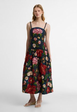 Γυναίκα που φορά αμάνικο, μεσομήκος φόρεμα σε σκούρο μπλε με μεγάλα κόκκινα, ροζ και λευκά λουλουδένια σχέδια και μπεζ πλατφόρμες, στέκεται σε λευκό φόντο.