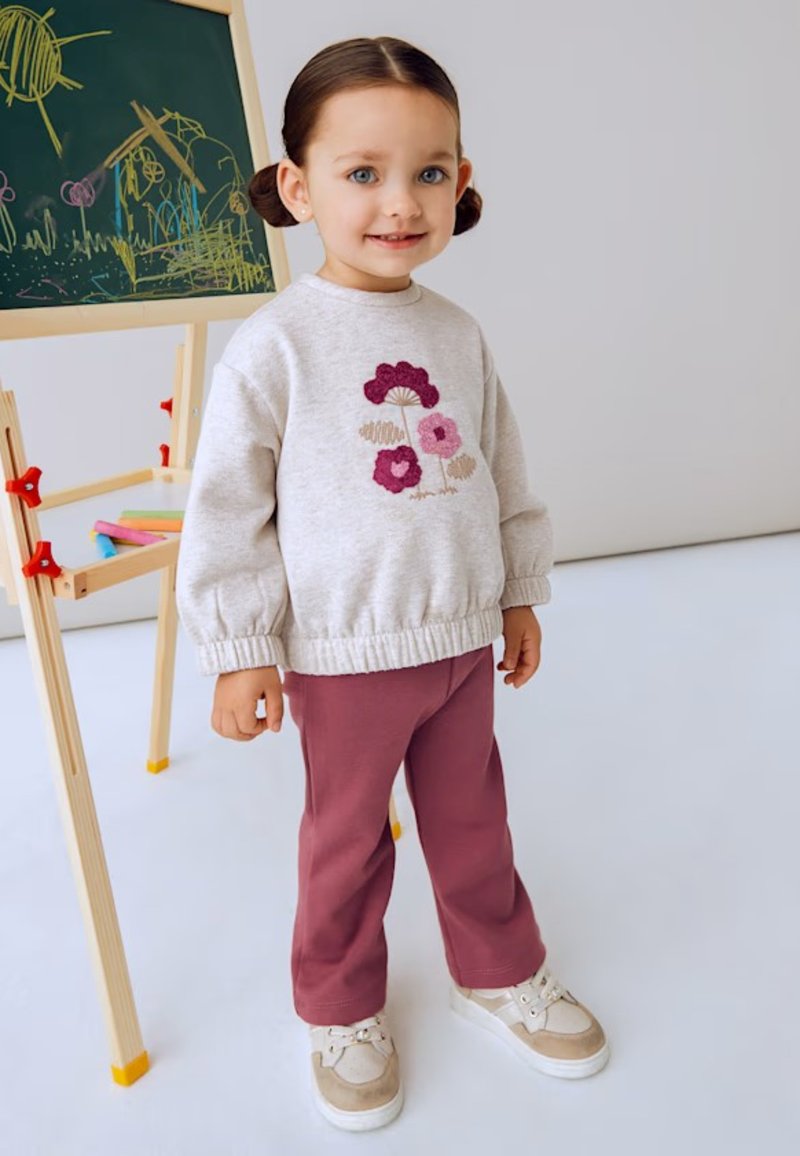 Maglione grigio chiaro con ricami floreali, abbinato a pantaloni rosa e sneaker bianche. Tessuto morbido e vestibilità comoda. Cavalletto in legno sullo sfondo.