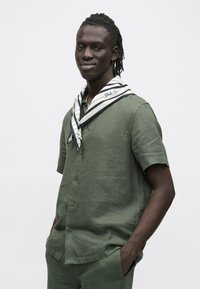 Camisa verde de linho de mangas curtas com frente de botões, complementada por um lenço branco e azul-marinho às riscas com detalhe de texto.