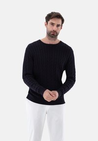 Dunkelblaues Kabelstrickpullover mit Rundhalsausschnitt und gerippten Bündchen, kombiniert mit weißen Hosen, vor einem schlichten hellen Hintergrund präsentiert.