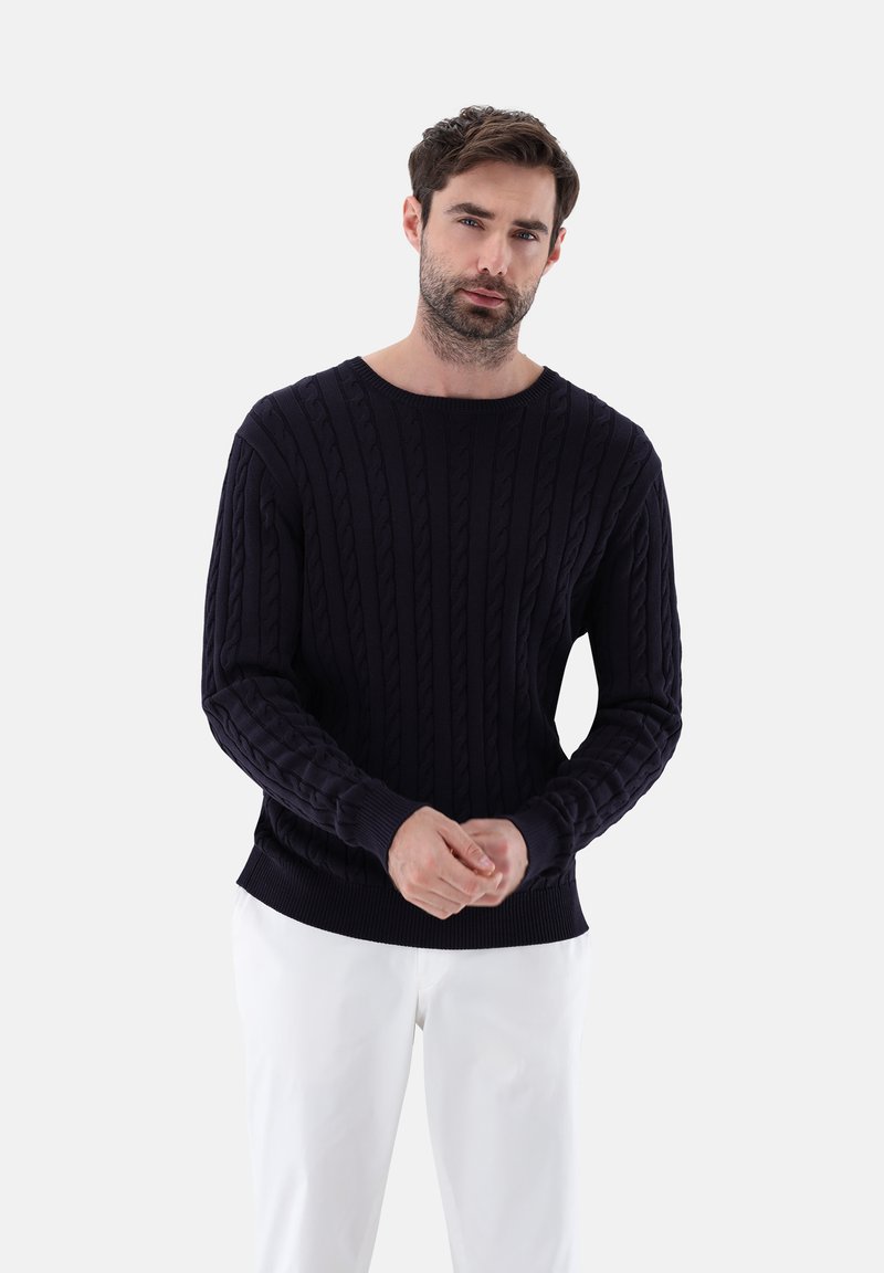 Dunkelblaues Kabelstrickpullover mit Rundhalsausschnitt und gerippten Bündchen, kombiniert mit weißen Hosen, vor einem schlichten hellen Hintergrund präsentiert.