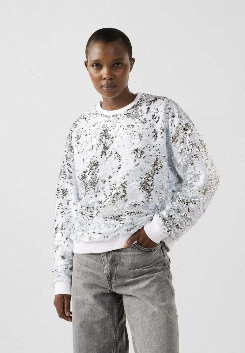 Persoon met kort haar die een zilveren pailletten trui met lange mouwen en vervaagde grijze jeans draagt, staande tegen een effen lichte achtergrond.