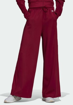 Persoon die losse bordeauxrode wijde broek met trekkoord in de taille en zijzakken draagt, en witte sneakers, staand met handen in de zakken.