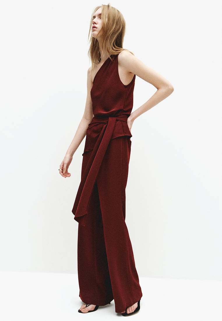 Femme aux cheveux clairs portant une combinaison sans manches, asymétrique sur une épaule, de couleur rouge foncé, avec des jambes larges et une longue ceinture à la taille, debout devant un fond blanc uni.