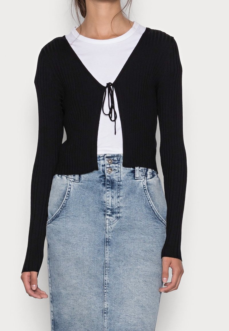 Femme portant un cardigan court noir côtelé à manches longues noué sur la poitrine, par-dessus une chemise blanche et une jupe en jean bleu clair taille haute.
