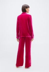 Γυναίκα που στέκεται ξυπόλητη φορώντας ένα σετ πυτζάμες από βελούδο σε έντονο ροζ χρώμα, με την πλάτη προς τον φακό, μπροστά σε απλό φωτεινό φόντο.