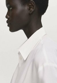 White collared shirt with a smooth texture, featuring a front button closure, shown on a model from a side profile against a neutral background.