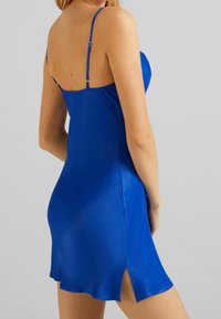 Woman wearing a short, bright blue satin slip dress with thin adjustable straps and a small side slit, against a plain light background.