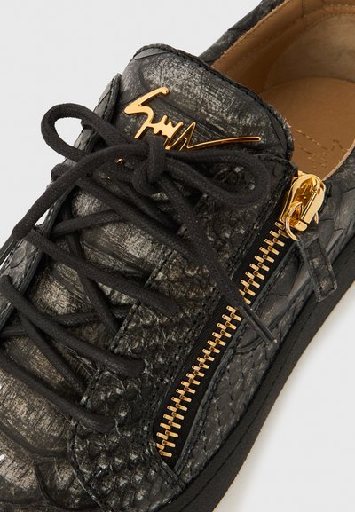 Svart texturerad lädersneaker med svarta skosnören, en guldfärgad dragkedja på sidan och en guldfärgad signaturdetalj på tungan.