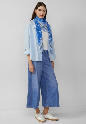 Jonge vrouw staat en draagt een lichtblauw overhemd, witte top, wijde blauwe spijkerbroek, witte sneakers en een blauw sjaaltje met patroon tegen een effen achtergrond.