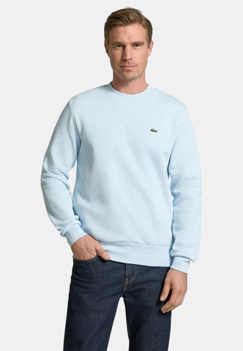 Sudadera de color azul claro en mezcla de algodón con cuello redondo acanalado y puños. Logo de Lacoste en verde oscuro en el lado izquierdo del pecho. Combinada con jeans oscuros.