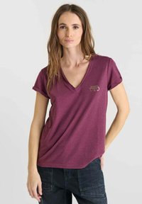 Vrouw met lang bruin haar draagt een plomverkleurige V-hals t-shirt met opgerolde mouwen en donkere broeken, staat met één hand in de zak tegen een effen achtergrond.