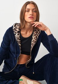 Námořnický sametový teplákový komplet s podšívkou kapuce v leopardím vzoru, zipovým předním dílem a bočními kapsami. Černý crop top uvnitř. Viditelné ležérní obuv.