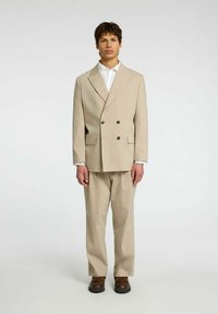 Beżowy dwurzędowy garnitur z wyłogowym kołnierzem, subtelny wzór w prążki, zestawiony z białą koszulą i brązowymi butami.