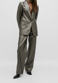 Donna che indossa un blazer oversize grigio metallico e pantaloni a gamba larga abbinati a stivaletti neri con tacco a spillo su uno sfondo neutro.