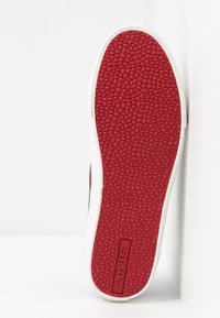 Semelle rouge en caoutchouc d'une basket avec surface texturée et bords blancs, arborant le logo Levi's embossé sur le côté. Design épuré et minimaliste.