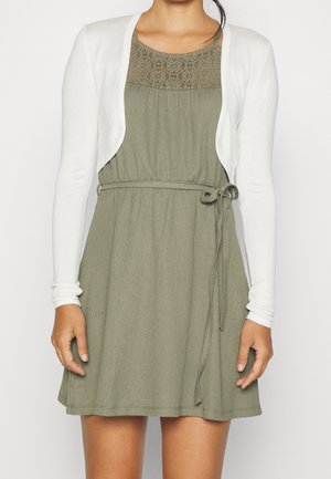 Robe vert olive avec une taille froncée et un lien, ornée de détails en dentelle au niveau de l'encolure, associée à un cardigan blanc ouvert sur le devant.