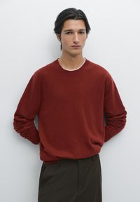 Rode kasjmier trui met een ronde hals, geribbelde manchetten en een ontspannen pasvorm. Draagt een donkere broek, geplaatst tegen een effen witte achtergrond.