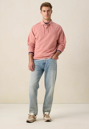 Lichtroze poloshirt met lange mouwen en een kraag, gecombineerd met lichtblauwe denim jeans. De schoenen zijn beige sneakers met rode accenten.