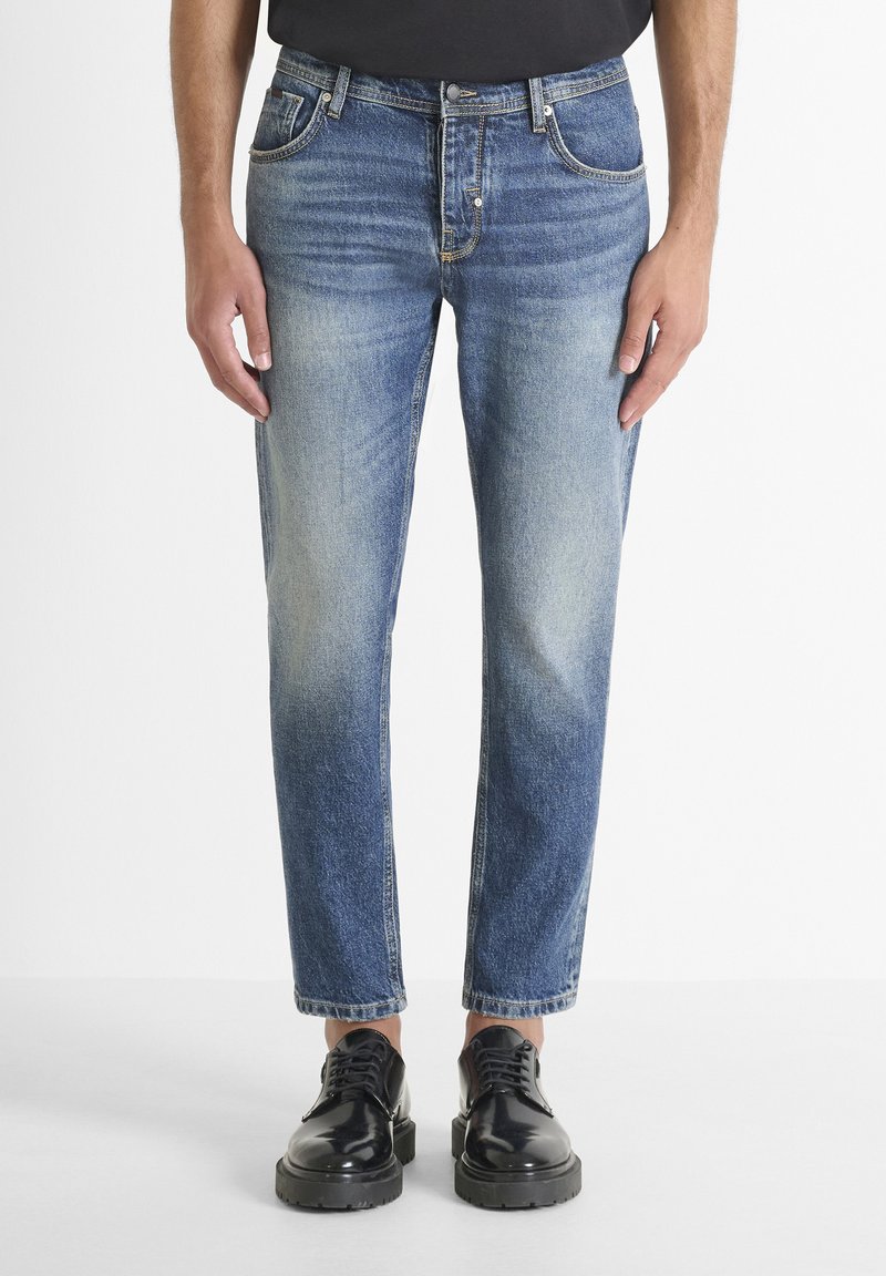 Jeans de mezclilla azul deslavado con un corte recto, diseño de cinco bolsillos y costuras visibles. Llevados con zapatos negros brillantes de cordones.
