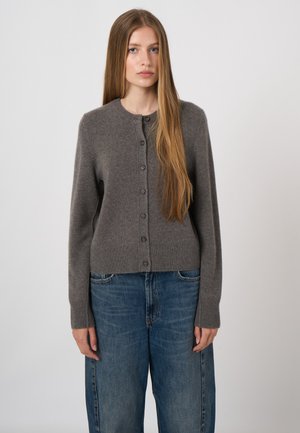 Jonge vrouw met lang, stijl haar, gekleed in een grijze gebuttonede cardigan en losse blauwe jeans, staand tegen een effen achtergrond.