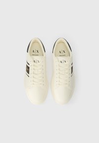 Kremne teniske z črnimi detajli, teksturiran zgornji material, ravni vezalni trakovi in logotip blagovne znamke natisnjen na ježku in straneh.