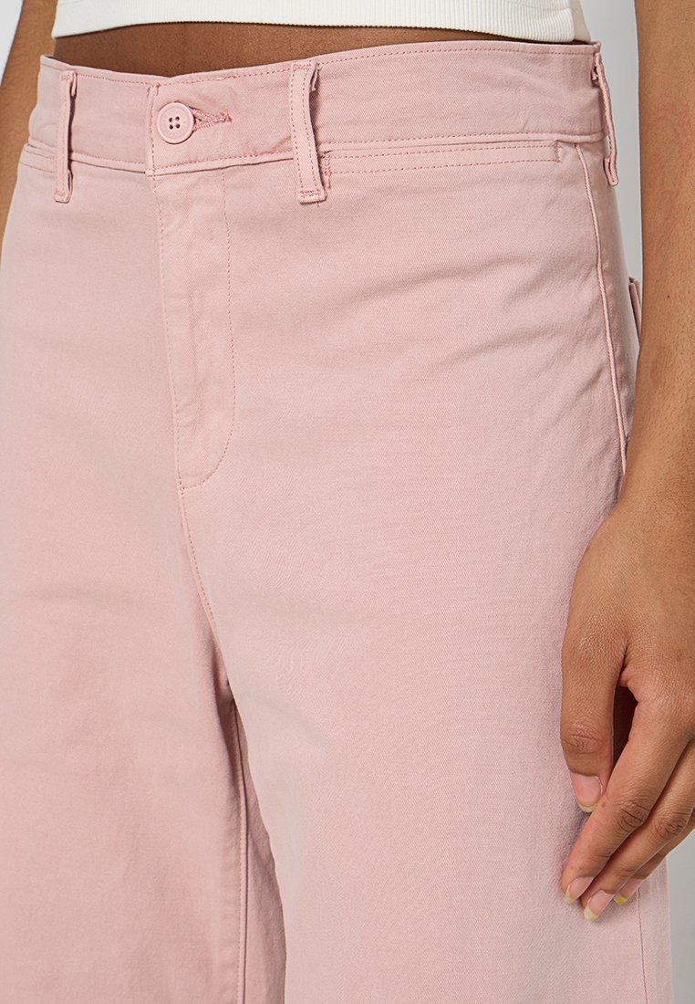 Pantalon en coton rose clair avec un design classique à cinq poches, fermeture à bouton et détails de couture visibles. Texture douce et coupe ajustée.