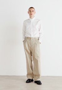 Chemise blanche à manches longues avec un pantalon plissé beige clair et des chaussures noires, sur fond blanc uni.