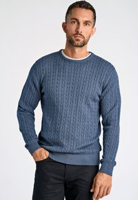 Blauer Kabel-strickpullover mit rundem Ausschnitt und gerippten Bündchen. Weiche Textur, mittleres Gewicht und taillierte Passform. Kombiniert mit dunklen Hosen.