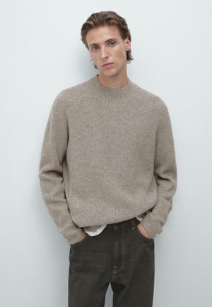 Jasnobrązowy sweter o prążkowanej fakturze z okrągłym dekoltem. Model ma na sobie ciemne spodnie; ręce w kieszeniach, na neutralnym tle.