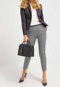 Zwarte getailleerde blazer over een witte blouse, gecombineerd met een strakke gebloemde broek en marineblauwe hoge hakken. Een gestructureerde zwarte handtas wordt in één hand vastgehouden.