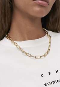 Guld kædedesign med rektangulære led og en poleret overflade, båret over en hvid skjorte med trykt tekst.